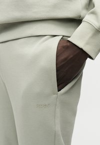 Χέρι με σκουρόχρωμο δέρμα που βρίσκεται μερικώς στην τσέπη ανοιχτόχρωμου γκρι φόρμας με ριμπ ζώνη μέσης και ανάγλυφο λογότυπο "HUGO".