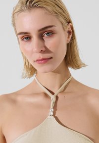 Beige Haltertop mit glänzender Textur, ausgestattet mit einem geknoteten Träger und drei dekorativen Perlen am Ausschnitt, vor einem hellen Hintergrund.