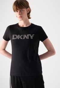 Črna bombažna majica s kratkimi rokavi, ki v središču prikazuje logotip "DKNY" okrašen s kristali. Klasičen kroj s plitkim izrezom in kratkimi rokavi.