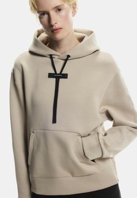 Bézs színű kapucnis pulóver elülső zsebbel, fekete húzózsinórral és logóval. Az anyag sima tapintású, és laza fazonú.