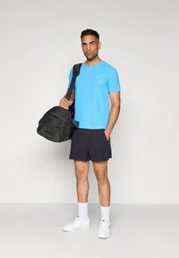 Mand stående iført lyseblå T-shirt, sorte shorts, hvide sokker og sneakers, med sort træningstaske over skulderen mod hvid baggrund.