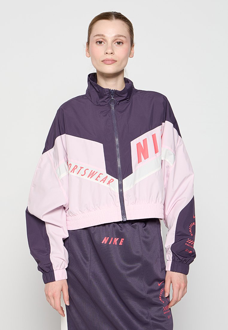 Joven mujer vistiendo una chaqueta deportiva Nike de color púrpura y rosa y una falda a juego con logotipos, de pie contra un fondo claro y liso.