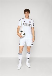 Uniforme da calcio bianco con accenti neri e dorati, caratterizzato dal logo del Real Madrid e il testo "Emirates Fly Better". Il giocatore tiene in mano un pallone con stelle nere.