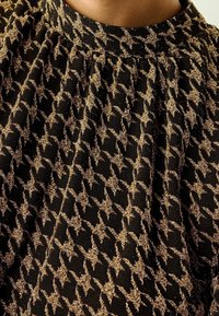 Tissu noir avec un motif étoile doré texturé, présentant des plis et un col montant. Le matériau a un léger éclat.