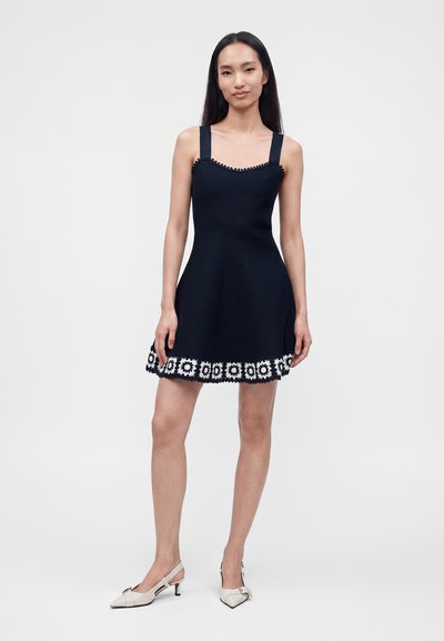 Femme debout portant une robe noire sans manches avec une bordure florale blanche à l'ourlet et des chaussures beiges à talons bas sur un fond uni.