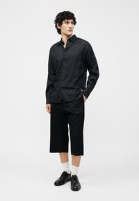 Άνδρας με σγουρά μαλλιά που φοράει μαύρο πουκάμισο με σχέδια και μακριά μανίκια, μαύρο παντελόνι με φαρδιές cropped μπατζάκια, λευκές κάλτσες και μαύρα επίσημα παπούτσια.