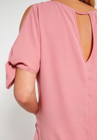 Růžový top s krátkým rukávem a kulatým výstřihem, který má odhalená záda. Materiál je hladký a lehký s mírným leskem.