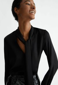 Blusa nera con dettaglio di lunga sciarpa plissettata legata al collo. Presenta una scollatura a V profonda e maniche lunghe e aderenti. Texture liscia.