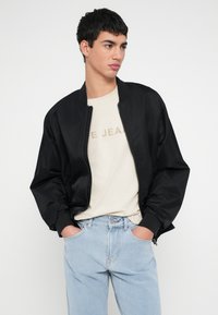 Must nahktagi, millel on sile tekstuur, soonilised varrukasääred ja esiosal lukk, kombineeritud heleda beežika t-särgiga ja helesiniste teksadega.