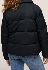 Sort pufferjakke med en quiltet design, der har en hætte og en kortet snit. Båret over blå denimjeans. Tekstureret stof med subtil glans.