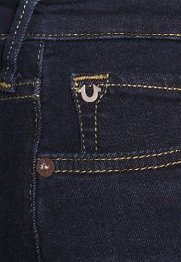 Dark denim jeans featuring a small front pocket, gold stitching, and a silver logo accent near the pocket. Textured fabric.