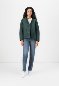 Veste matelassée vert foncé avec des boutons-pression, deux poches avant, associée à un jean bleu clair et des baskets blanches, sur un fond uni.