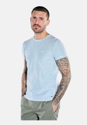 Mann mit kurzen Haaren und Tätowierungen an beiden Armen, trägt ein hellblaues T-Shirt und grüne Hose, die Hände in den Taschen, neutraler Hintergrund.