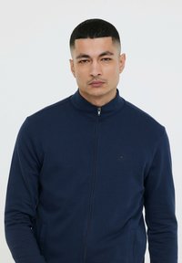 Veste zippée bleu marine en tissu lisse avec un col montant, comportant des poches latérales et un logo discret sur la poitrine.