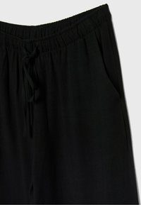 Pantalon ample noir avec taille élastique et cordon, avec une poche latérale sur un fond clair.