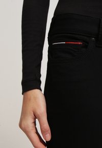 Pantalones de mezclilla negros con un bolsillo frontal que presenta acentos de costura en rojo, blanco y azul. Textura suave y diseño ajustado visibles.