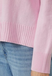 Hellrosa Pullover mit geripptem Saum und Bündchen, hergestellt aus weichem Strickmaterial. Kombiniert mit blauen Jeans.