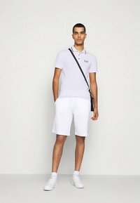 Bílá polokošile s límečkem, krátkými rukávy a logem. Kombinována s bílými šortkami a bílými teniskami. Černá kabelka s popruhem přes tělo nošená napříč tělem.