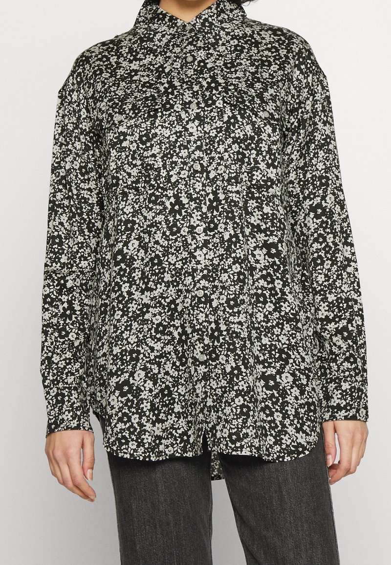 Chemise à manches longues à boutons, noire avec imprimé floral blanc. Dotée d'un col et d'une coupe ample. Le tissu semble soyeux avec une texture lisse.