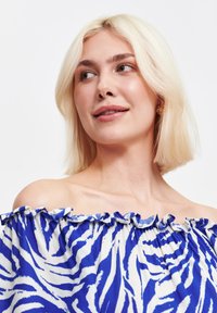 Blondynka z lekkim uśmiechem, nosząca bluzkę na ramiączkach z niebiesko-białym wzorem zebry, zwrócona lekko w prawo.