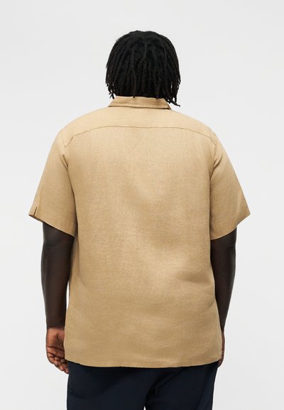 Loom tumeda nahaga ja dreadlockidega, seljas lahtine lühikeste varrukatega beež triiksärk, selja poolt vaadatuna tavalise tausta ees.