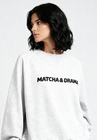 Felpa grigio chiaro con scritta nera "MATCHA & DRAMA", collo rotondo e maniche raglan, realizzata in un tessuto morbido e strutturato.