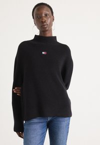 Svart strikket genser med høy krage, med en liten flerfarget logo på brystet. Lange ermer og en avslappet passform. Brukt med blå jeans.