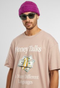 Tricou roz deschis cu textul "Money Talks" și o grafică cu bani în stivă. Modelul poartă ochelari de soare verzi și o căciulă tricotată roz.