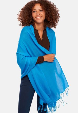 Châle bleu en tissu doux et léger avec des bords frangés, drapé sur un haut marron, porté par une femme aux cheveux bouclés.