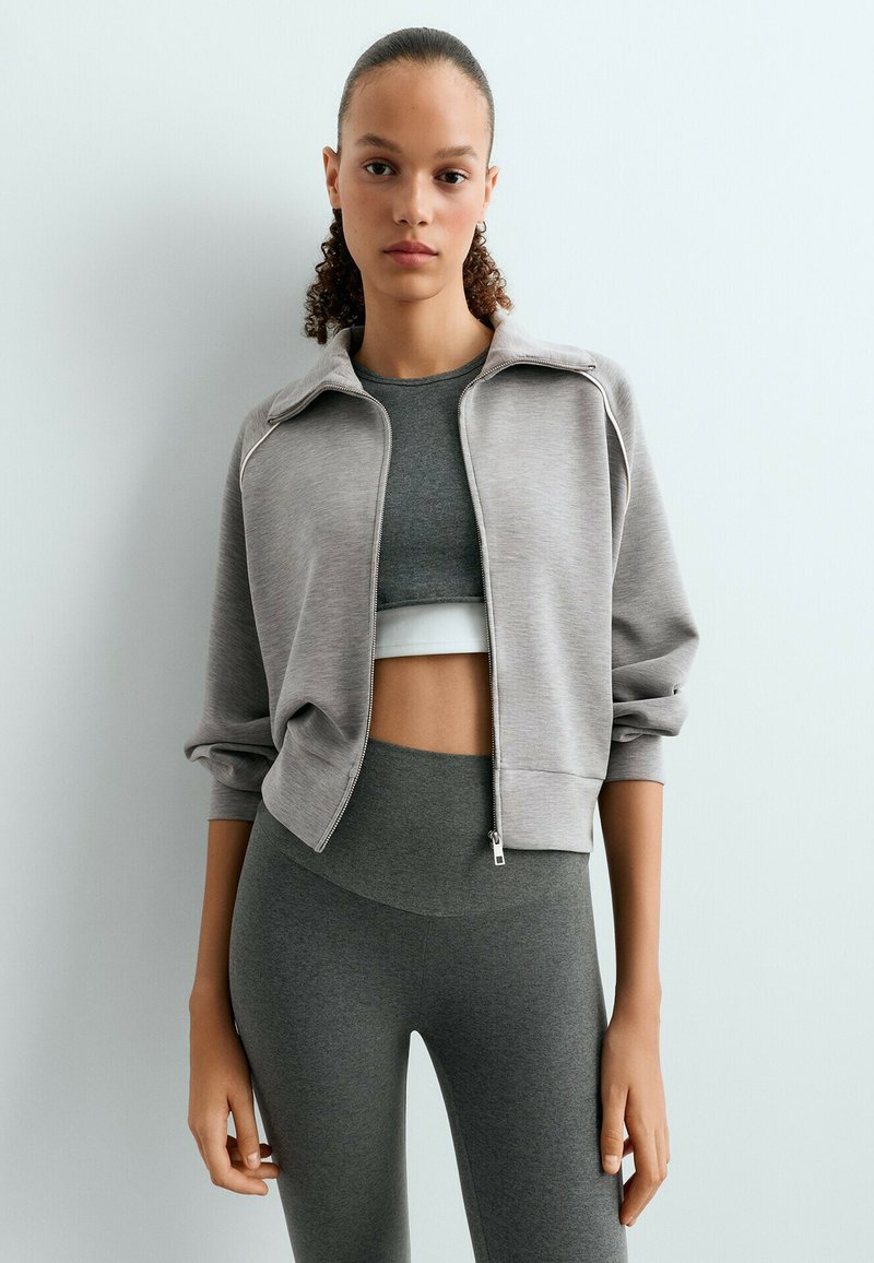 Jeune femme aux cheveux bouclés vêtue d'une veste zippée grise par-dessus un crop top gris foncé et un legging assorti, sur un fond clair uni.