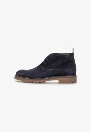 Stivaletto alla caviglia in camoscio nero con suola in gomma marrone con tacchetti e lacci blu scuro, mostrato di profilo su sfondo bianco.