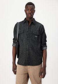Black denim shirt with button-front, two chest pockets, and rolled cuffs. Paired with light-colored pants. Simple textile design.