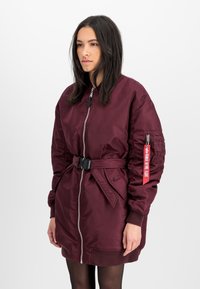 Burgundy bomber jacket with a front zipper, belted waist, ribbed cuffs, and a patch on the sleeve. Smooth, shiny fabric with a relaxed fit.