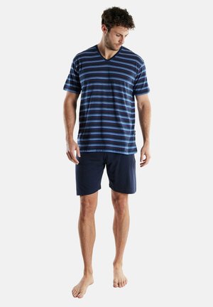 Man standing barefoot wearing navy blue shorts and a navy blue striped short-sleeve V-neck shirt, looking down to his left.