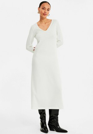 Langes, weißes, geripptes Kleid mit V-Ausschnitt und langen Ärmeln, kombiniert mit schwarzen kniehohen Stiefeletten mit spitzer Zehenpartie und Creolen.