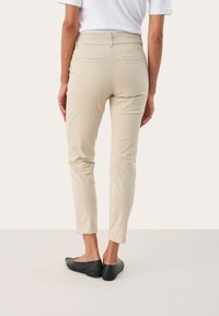 Pantalon slim beige clair avec une texture lisse, doté de poches arrière et d'une taille mi-haute, associé à des chaussures plates noires.