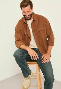 Braun- und orangefarbener karierter Hemd, hellbeige T-Shirt, dunkelblaue Jeans und beige Schuhe. Mann sitzt auf einem Holzschemel, die Hände auf den Beinen ruhend.