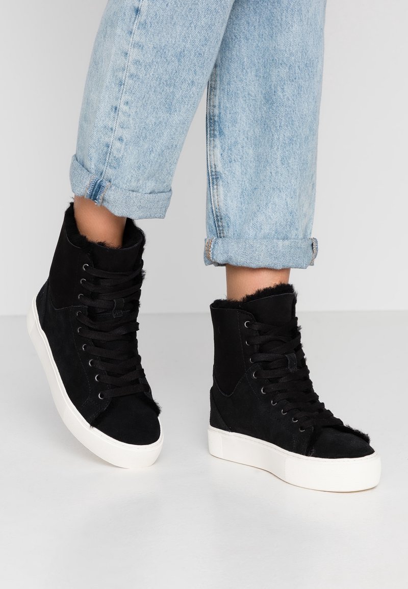 Personne portant un jean bleu clair retroussé et des baskets montantes noires avec des semelles blanches sur un fond gris clair.