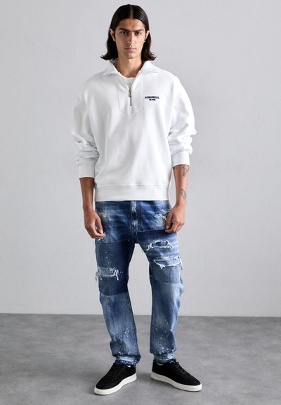 Vit sweatshirt med dragkedja i kragen, ihop med slitna blå denimjeans med lappar och fransiga detaljer. Svarta skor fulländar looken.