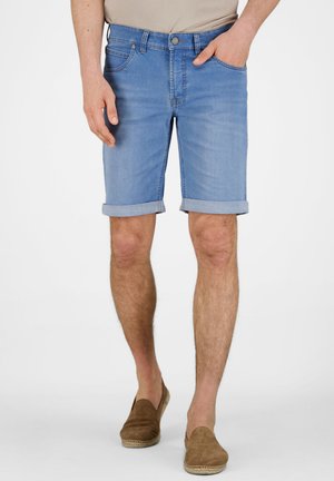 Mann trägt hellblaue hochgekrempelte Jeansshorts mit beigen Slippern und linker Hand in der Hosentasche, steht vor weißem Hintergrund.