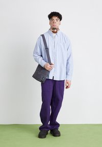 Vaatetus rento, jossa on vaaleansininen raidallinen napillinen paita, violetit leveät housut ja harmaa olkalaukku olkahihnalla.