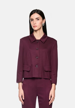 Woman with straight black hair wears a buttoned dark purple jacket with collar and matching pants against a plain light background.