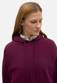 Sudadera morada de tejido texturizado, con capucha ajustable y mangas largas, llevada sobre una camisa blanca con cuello.