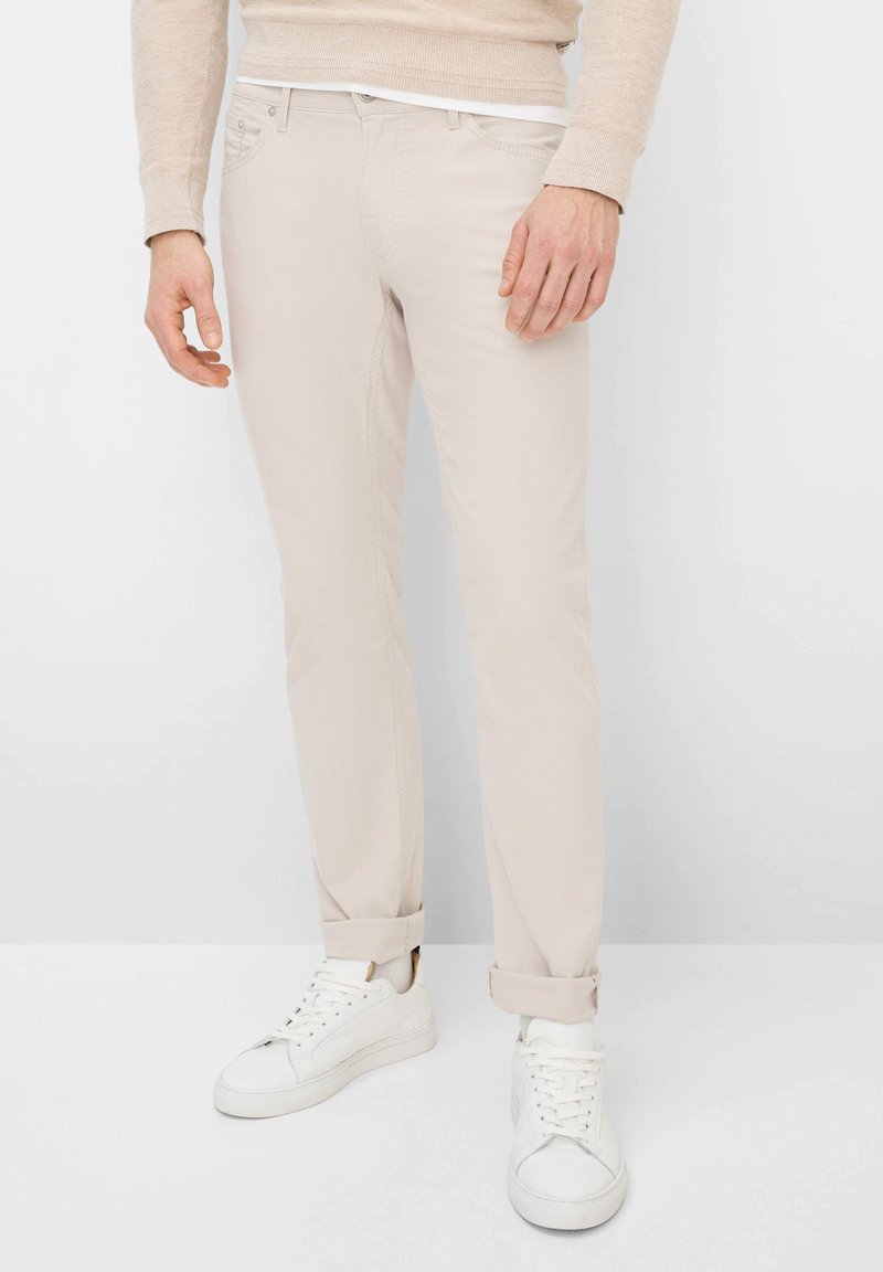 Mann trägt beige Chinos, weiße Turnschuhe und einen hellbeigen Pullover und steht vor einem weißen Hintergrund.