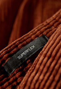 Bruine, gestructureerde stof met verticale ribbels en een zwart etiket met de tekst "SUPERFLEX" in het wit. Close-up details van het materiaal.