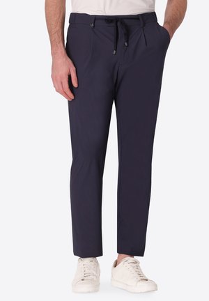 Uomo con pantaloni slim fit blu navy con vita con coulisse e sneakers bianche, in piedi con una mano in tasca.