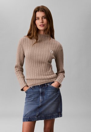 Kvinde med langt brunt hår iført en beige ribstrikket sweater og blå denim nederdel med hænderne i nederdelens lommer mod en grå baggrund.