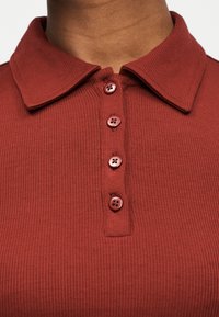 Κοντινή λήψη ενός ατόμου που φοράει πόλο μπλούζα σε τούβλινο κόκκινο χρώμα με ριμπ υφή, τέσσερα κουμπιά στο ί διο χρώμα και διπλωμένο γιακά.