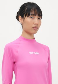 Rash guard dugih rukava u svijetlo ružičastoj boji, napravljen od rastezljivog materijala, s bijelim "RIP CURL" logotipom na prsima i ravnim šavovima.