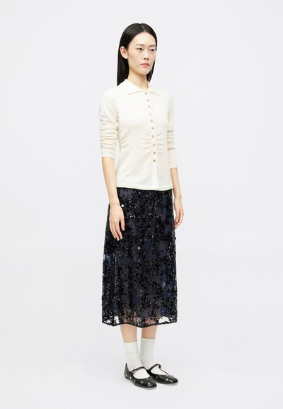Cream, button-down, long-sleeve top with gathered front. Navy beaded, sheer midi skirt, white socks, black shoes with metal detailing.